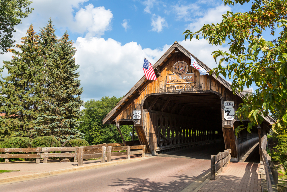 The entrance to the Holz-Brucke covered wooden bridge in the small town of Frankenmuth Michigan. It has a Dutch style sign on it and is surrounded by trees with green leaves. 