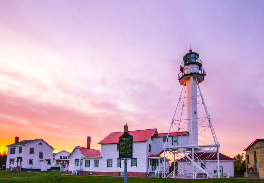 The exterior of the Great Lakes Shipwreck Museum. It is several old buildings, all painted white. They have red rooves and black window trims. There is a white lighthouse with black accents. It is sunset and the sky is pink and purple. 