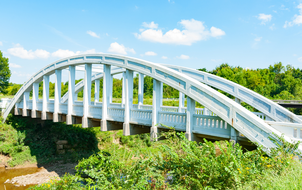 A bluish-white bridge that has an arch shape on both sides of the road. There is a river or stream running underneath it. Around the bridge there is a ton of greenery like shrubs, grasses, and trees