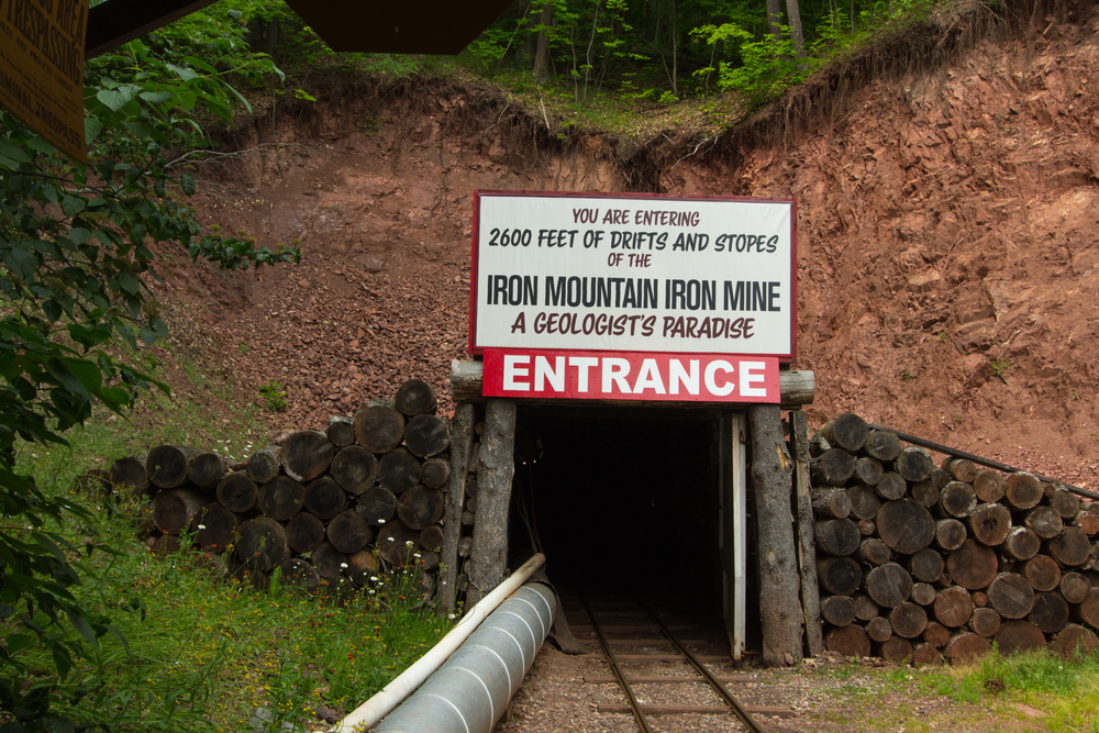 The entrance to the Iron Mountain Iron Mine. It is a dark tunnel with wood blocks around it and a large white sign that says 'you are entering 2600 feet of drifts and stopes of the iron mountain iron min a geologist's paradise' with a red Entrance sign under that. The tunnel is in the side of a rocky hill with grass in front of it. Its one of the best things to do in the UP.