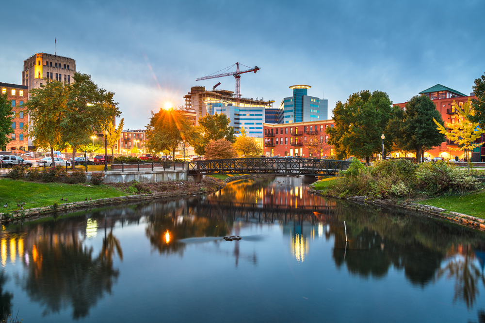 The city skyline of Kalamazoo Michigan, a large city. It is twilight, so the city is all lit up and the sun is setting. The main part of the picture is a large pond in the middle of the city with a metal bridge going across it with the skyline behind it. Its a great stop for Michigan road trips