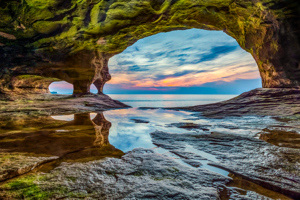 The view of Lake Superior from a rock cave. The rock cave has lots of levels in in it and there is water reflecting the cave and sky. The rocks in the cave are tan, green, red, and orange. Things to do in the UP