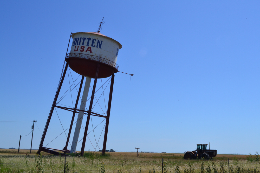 A white water tower leaning over at a serious angle over a field. In the field, a tractor is parked near the leaning tower and there is grass growing. 