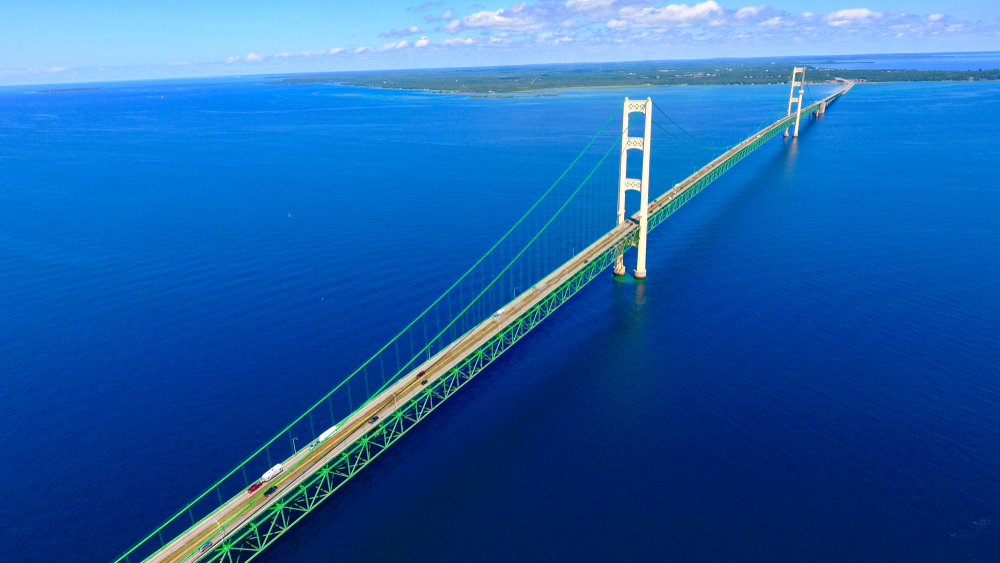 An aerial view of the Mackinac Bridge over the peaceful lake water. The bridge is similar in style to the golden gate bridge but it is painted white and green. It leads in this photo to Mackinac Island. 