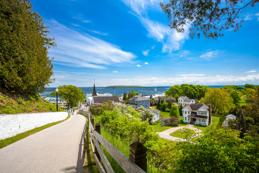 The view from a walking trail of Mackinac Island. You can see historic homes, a church steeple, and the lake. There are a ton of trees and tall grasses near the walking trail and throughout the view of the island. Mackinac Island are great things to do in the UP