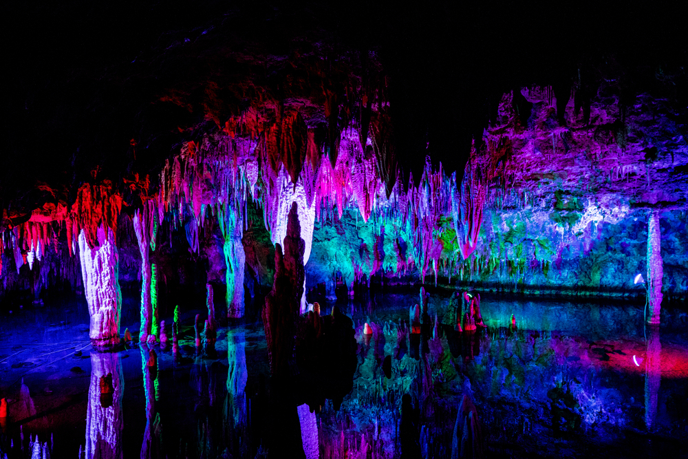 The interior of a large cavern system in Missouri. The stalagmites and stalactites are lit up using red, purple, green, pink, and blue lights. It looks like there is water at the bottom that is reflecting some of the lights further