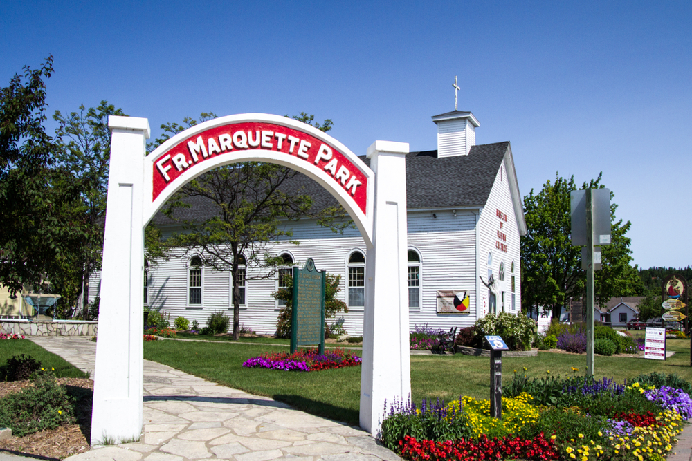 The entrance to the Father Marquette Park at the Ojibwa Cultural Museum. There is an arch entrance to the park that is white with a red sign that has the name on it. Near it there are red, yellow, and purple flowers. There is also an old church that is painted white behind the park entrance sign. things to do in the UP