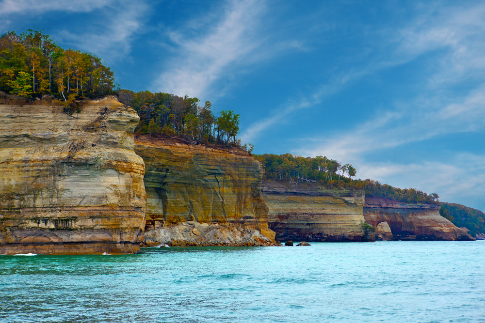 A view of the various cliffs at the Pictured Rocks National Lakeshore. The cliffs have layers of different colored rock, like yellow, green, and a rusty orange. There are trees on the rocks, most of them have green leaves but a few have orange leaves. It is a sunny day with a blue sky and the water looks very blue. Its one of the best stops on Michigan road trips