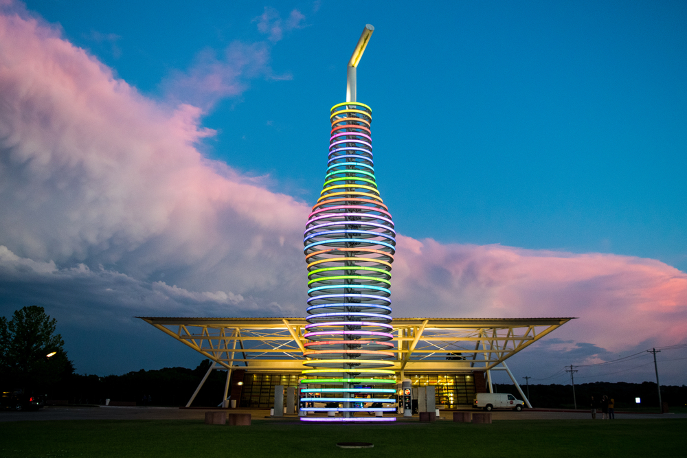 A large multi colored soda bottle. The bottle is colored with rings of lights. The lights are shades of yellow, orange, red, pink, purple, blue, and green. Behind the bottle there is a Mid Century style building with lights and windows and a van parked in front of it. It is sunset and there is a large swatch of pink and purple clouds in the background