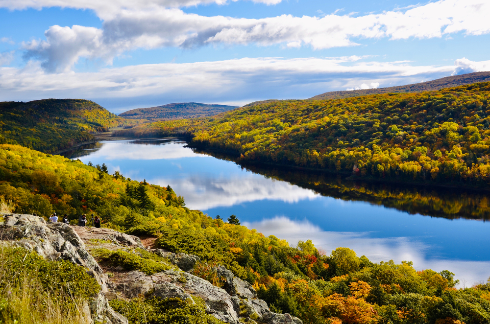 A large lake at the Porcupine Wilderness State Park. There are hills covered in trees and some of them have yellow and orange leaves. There are some rock formations that people are sitting on. The sunny partially cloudy sky is reflected in the lake.