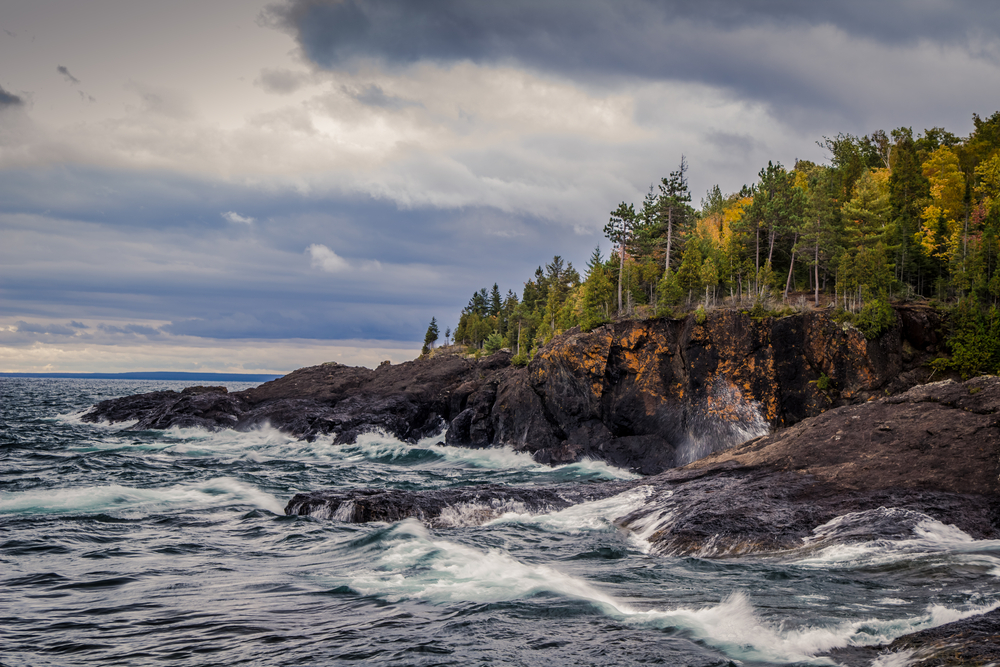 The rocky shores of Presque Isle Park on a cloudy day. There are waves crashing against the rocky shore. The rock formations are dark brown with some having orange flecks in them. Just pass the rocky shore is a landscape of trees with mostly green leaves, but a few of the trees have yellow leaves. 