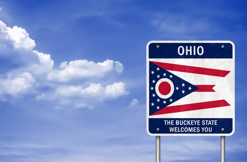 A road sign for welcoming you to Ohio. On the sign is the state flag, which is blue a triangle with stars and an O in it, and two white stripes and three red stripes. The sign says 'Ohio, the buckeye state welcomes you'. There is a pretty sunny sky with fluffy clouds behind the sign. Ohio road trips will always see this sign
