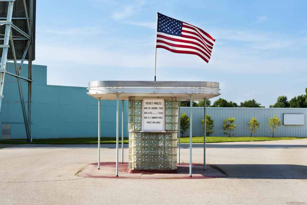 The ticket booth at a retro original route 66 drive in theater. It is made of glass bricks, has an overhang on all sides, and an American flag flying above it. It also has the ticket prices on the side on a white board with black lettering. In the background, you can see part of the drive in screen. One of the best route 66 attractions