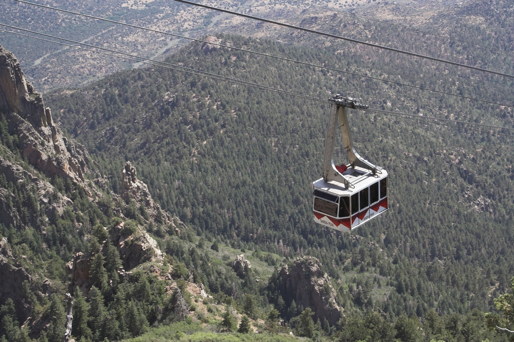 The Sandia Peak Tramway, which is a suspended shuttle over the side of the Sandia Peak Mountain in New Mexico. On the mountain there are trees and rocky cliffs Route 66 attractions.