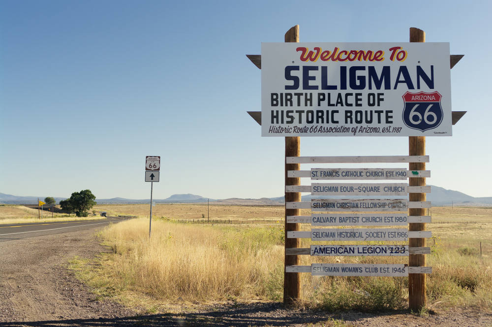 The Welcome sign when you enter Seligman Arizona. It is on the side of a road with tall grass around it and in the very far distance there are mountains. The sign has dark blue, yellow, and red lettering. Down the front it has information on each individual historic site in the town. One of the best route 66 roadside attractions