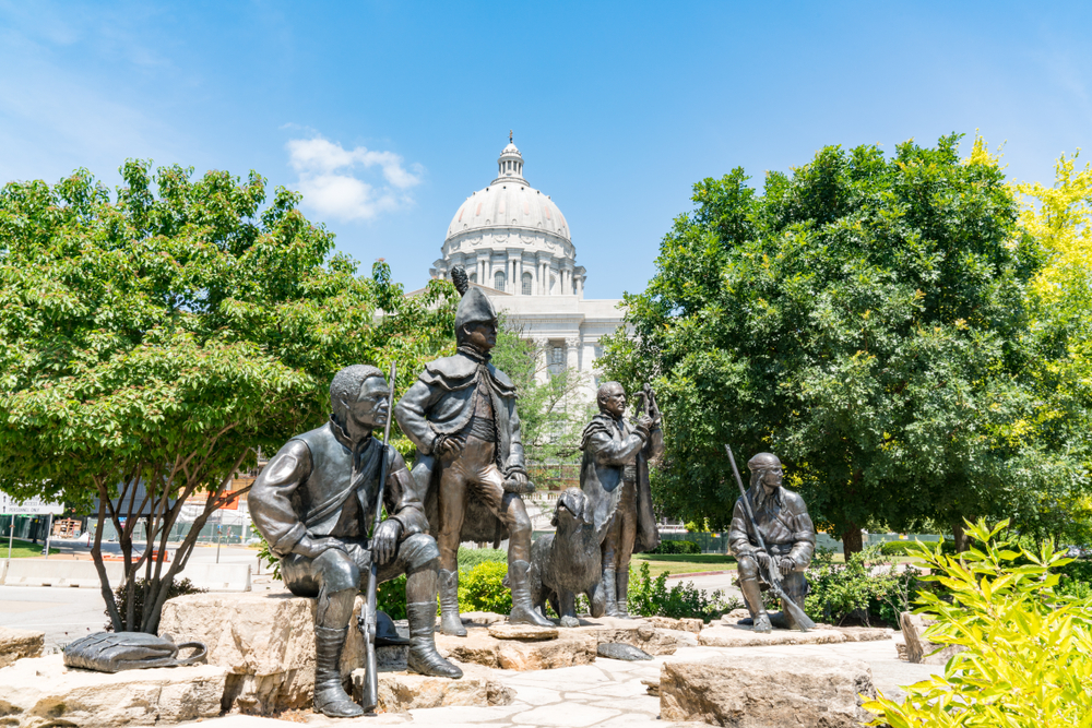 Bronze monument of 4 men and 1 dog on standing on stones with capital building in background. Things to do in Missouri/