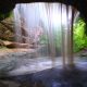 A cave with a waterfall flowing over the side and lush green trees behind. In an article about waterfalls in Illinois