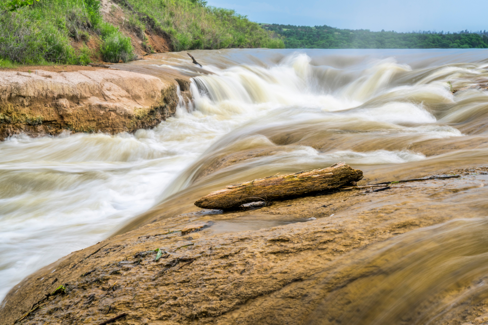 Water from calm river being pulled over rocks and churning below. Piece of wood in foreground.