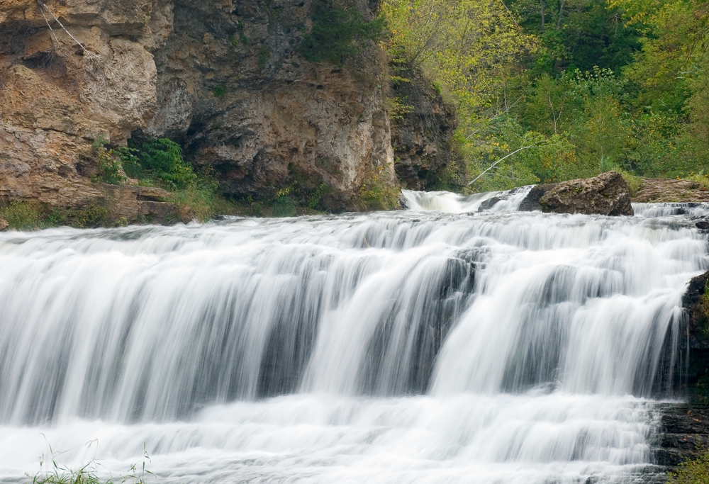 White raging waters flow over rock formations into waters below, with natural stone and green trees in background. Waterfalls in the Midwest.