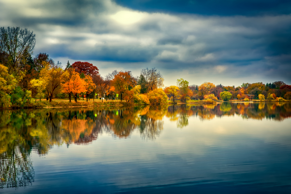 Looking out at a lake from the Bde Maka Ska North Beach. The trees around the lake are very colorful. They have red, orange, yellow, green, and brown leaves. The sky is very dark and cloudy and the trees and sky are almost perfectly reflected on the lake's surface. 