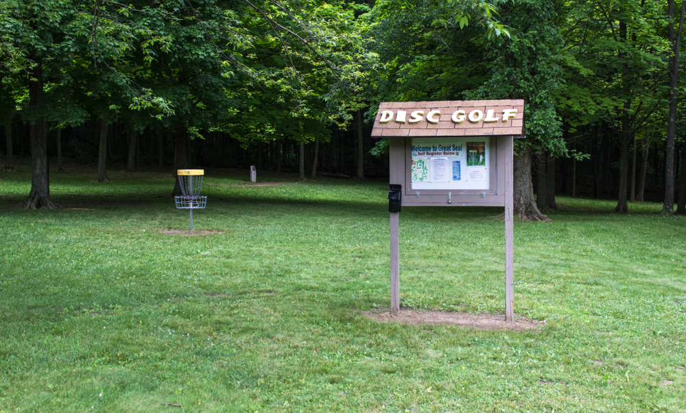 In this state park in Ohio, a sign and a disc golf cage are pictured amongst green grass and large green trees.  