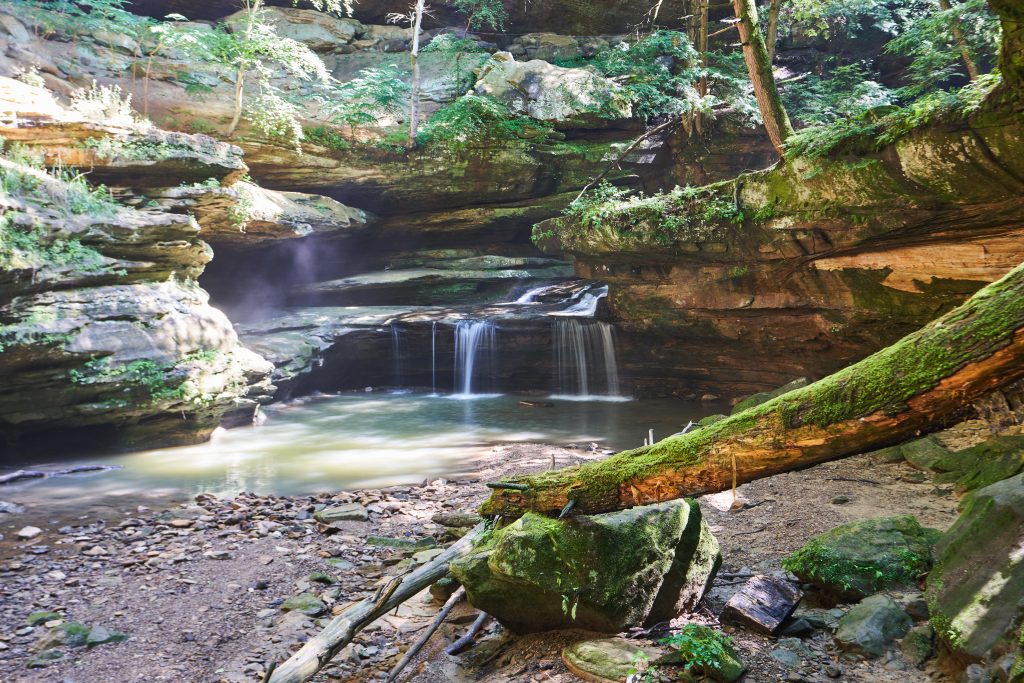 A waterfall is pictured cascading over mossy rocks in green, forest surroundings at Hocking Hills State Park.