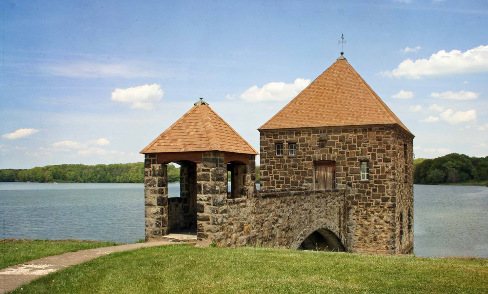 Brown stone structure with light brown pointed roofs with ornate arch leading to wooden front door, with lake in background. 