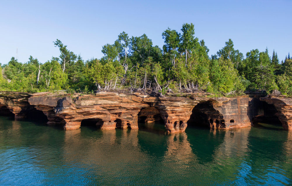 The unique rocky shore of the Apostle Island National Lakeshore. The shore has trees growing on it with green leaves. One of the most unique places for a Wisconsin weekend getaway.