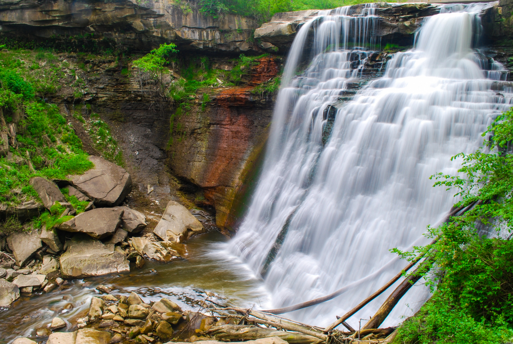 A large cascading waterfall flowing into a small river surrounded by rocks. There is grass and moss growing on the rocks and some of the rocks are multicolored in the Cuyahoga Valley National Park. 