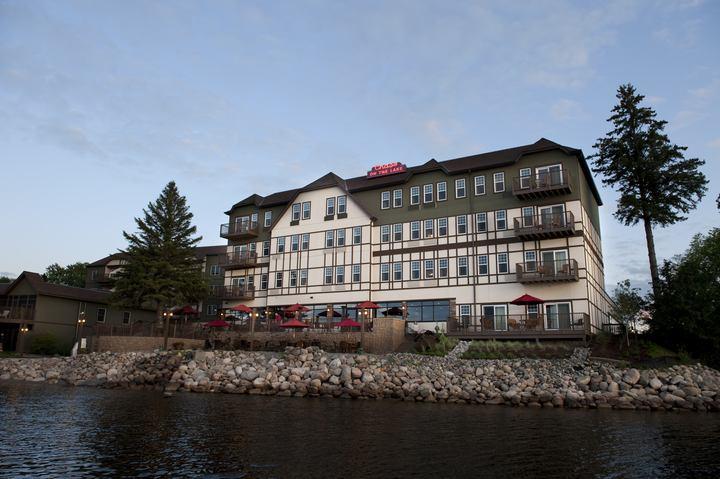 The exterior of an old style lake resort. It is white and dark green with lots of windows. There is a patio with umbrellas and is is right on the rocky shore of a lake. 