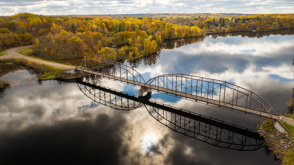 A bridge going over the Mississippi River to a shore of the river covered in trees. The trees have yellow, orange, and some green leaves. You can see clouds reflected in the water of the river. 