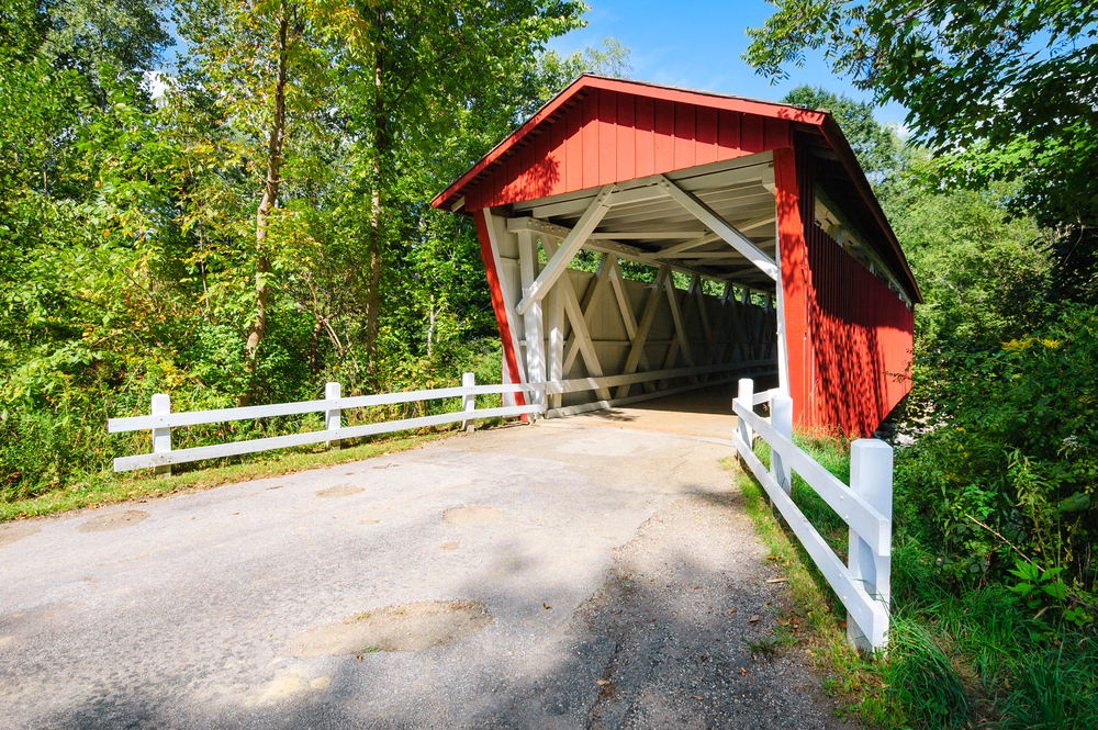 A road leading to a large red and white covered bridge. There is a white fence next to the road leading to the bridge. The area is surrounded by trees and it is a sunny day. 