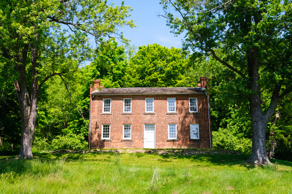 The front exterior of a brick house with lots of windows with white trim. There are two old trees on either side of the house, a grass front yard, and there are dense trees behind the house. 