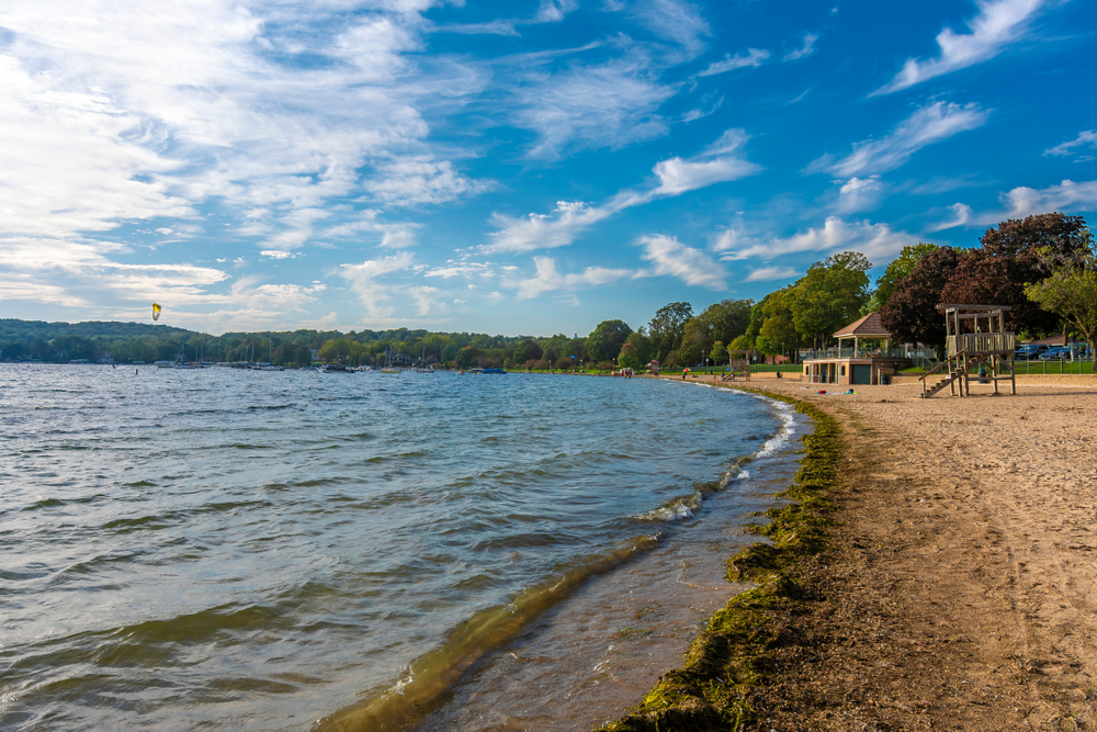 The shore of Lake Geneva on a sunny day. On the shore you can see a few playground structures and trees with green and brown leaves. 