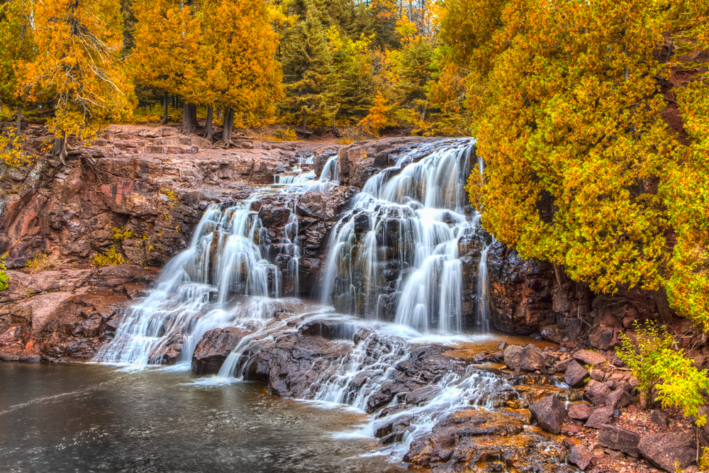 A large rocky waterfall with many splits in it. The cliff is very rocky and surrounded by trees. The trees all have yellow and orange leaves, with some of the leaves are still green. It is a beautiful fall in Minnesota view.