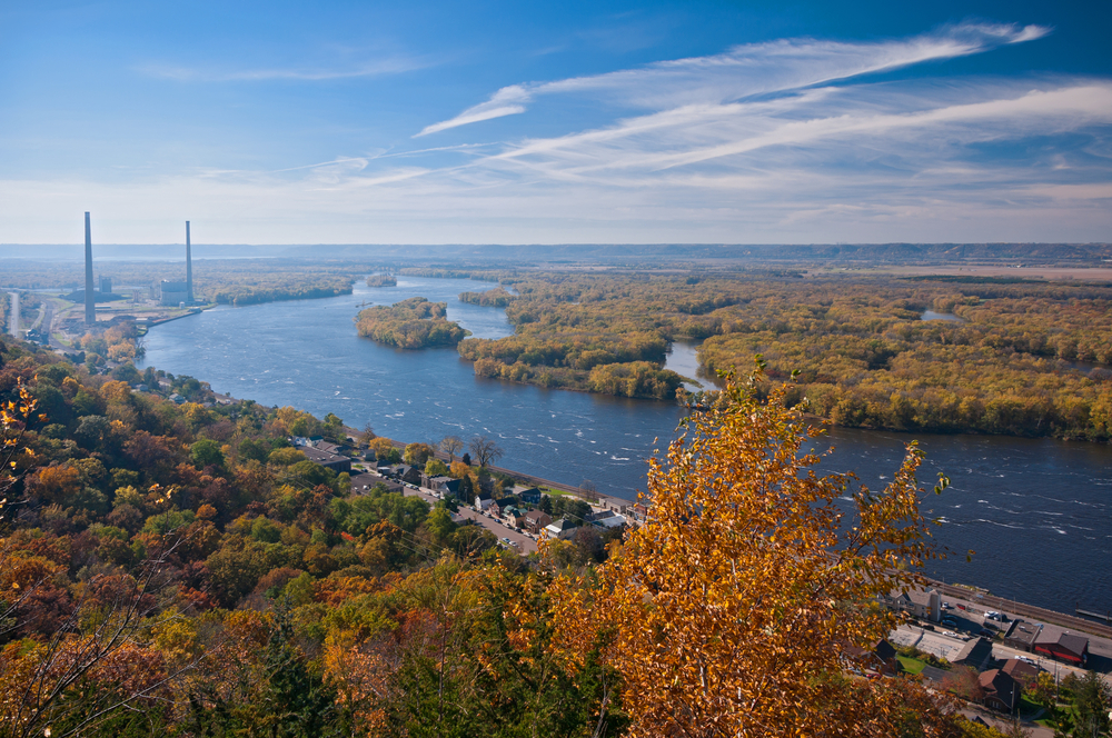 An aerial view of the Mississippi River with a town on the edge of the river. You can see lots of trees that are changing in the fall and have red, orange, yellow, and green leaves.