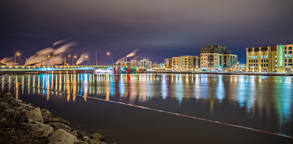 Looking across the river at the city of Green Bay Wisconsin all lit up at night. There are tall buildings, smoke stacks, and colored lights. 