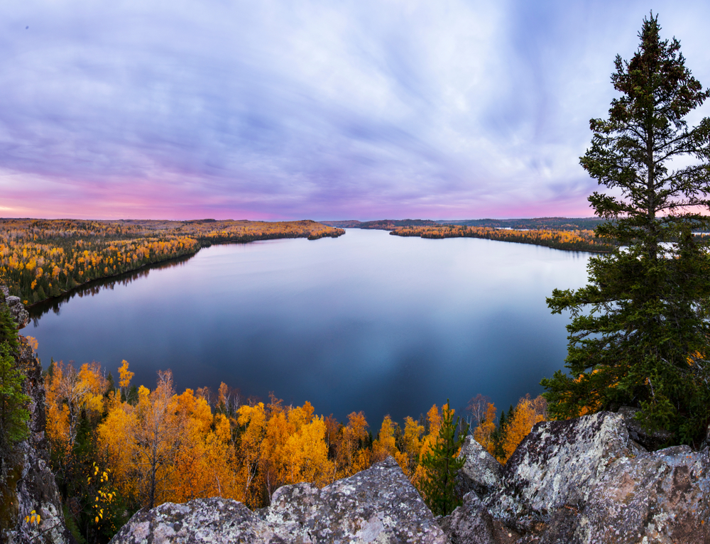 The view of Honeymoon Bluff in Superior National Forest. There is a rocky cliff with trees at the bottom and then a large body of water. On the shore of the body of water there are tons of trees. The trees have yellow, orange, and red leaves. Some of them still have green leaves. It is sunset, so the sky is blue, pink, and purple. It is a beautiful fall in Minnesota view