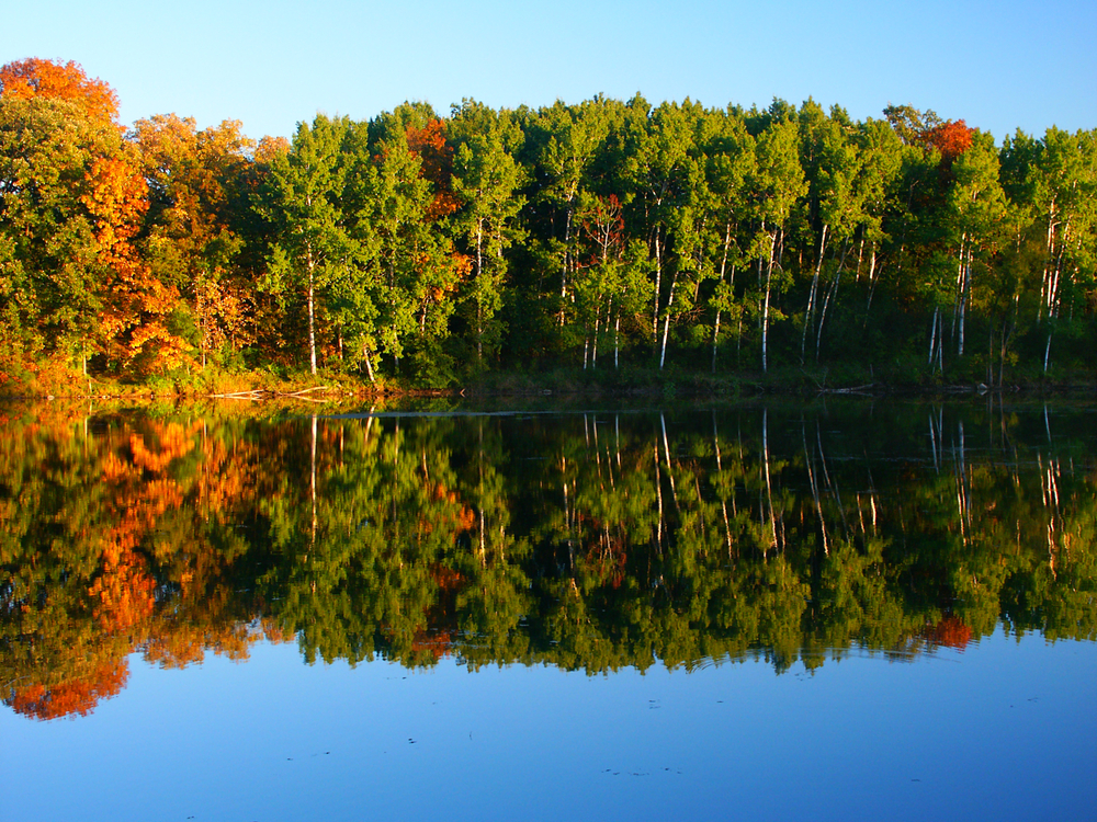 A lake with trees on the shore at Kettle Moraine State Park. The trees are mostly green, but a few have red, orange, and yellow leaves. The trees are reflected in the water of the lake. 