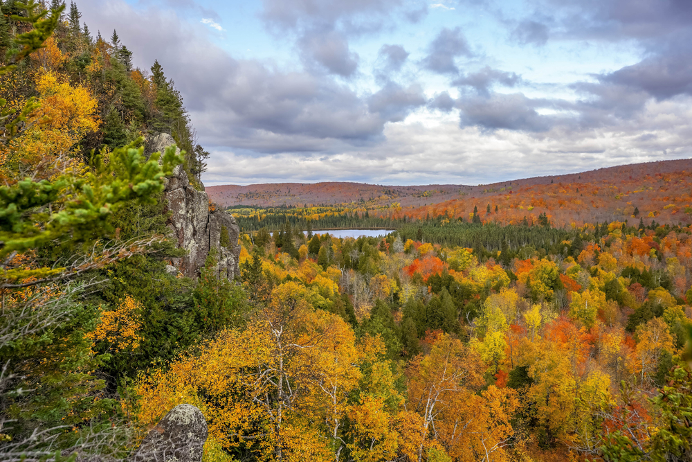 The view of the Superior National Forest from Oberg Mountain. There is a rock formation on the left side of the view. You can see hills covered in trees and a partial view of a body of water. The trees have green, brown, yellow, orange, and red leaves. Its a great Fall in Minnesota view. 