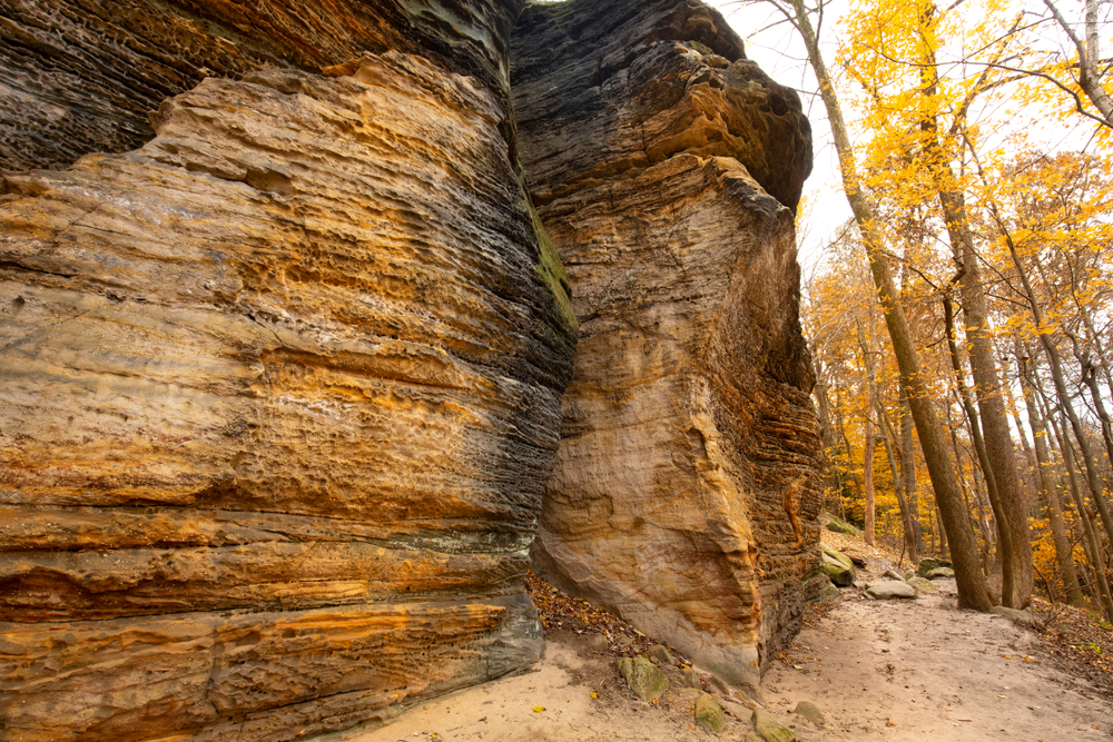 Large rock formations on the side of a trail that are multicolored. The other side of the narrow trail is surrounded by trees. 