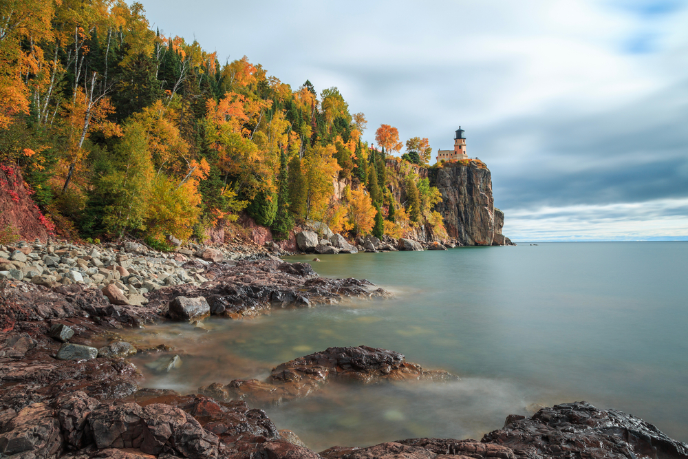 The view from the shore of a lake of the Split Rock Lighthouse. The lighthouse is perched on a rocky cliff and looks to be a peachy color and black. Behind it is a dense forest full of trees that come to the rocky shore line. The trees have yellow, orange, and green leaves. The sky is cloudy.