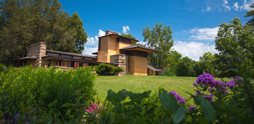 Looking through bushes with purple flowers to see a landscaped yard with a angular home designed by Frank Lloyd Wright. The home is pale yellow stucco with brown stone and a brown roof. The home is surround by trees.