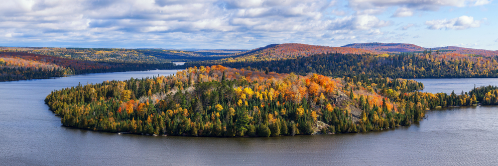 The view of the landscape at Superior National Forest. There is a large body of water surrounded large mountains and hills. The landscape is full of dense forest and goes on for miles. The trees in the forest are green, yellow, red, and orange. The sky is light blue with big fluffy clouds