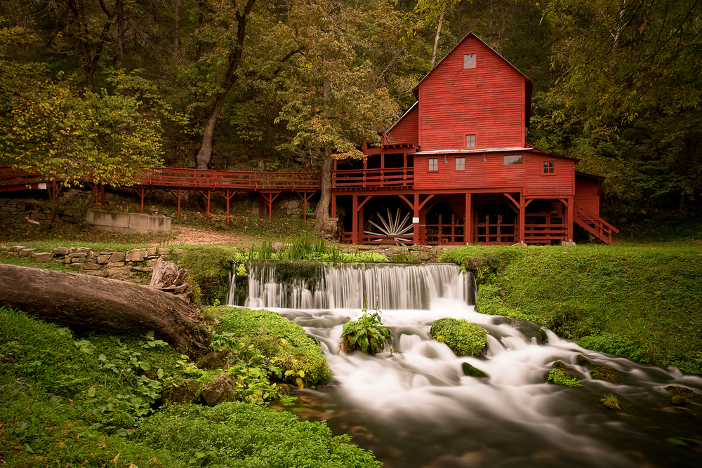 Rural red grist mill with wide waterfall surrounded by green trees.