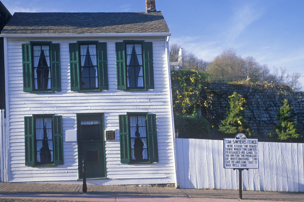 White wood-plank house with green shutters, white fence with historical marker in front of it. Things to do in Missouri.