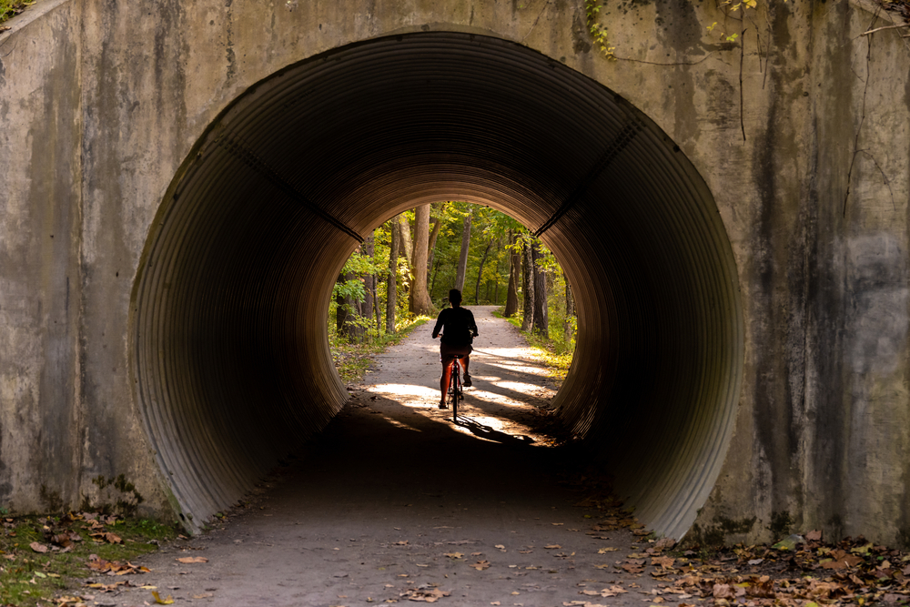A circular tunnel in concrete under a bridge that has a wide path in it. Someone is biking down the path towards a part that is lined with trees with green leaves. 