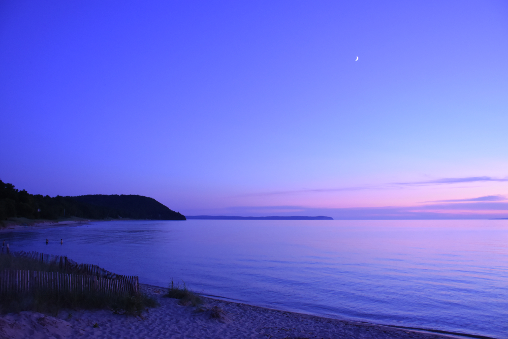 Beautiful purple sky and  calm waters on Michigan beach, with sand and grassy hills in foreground.