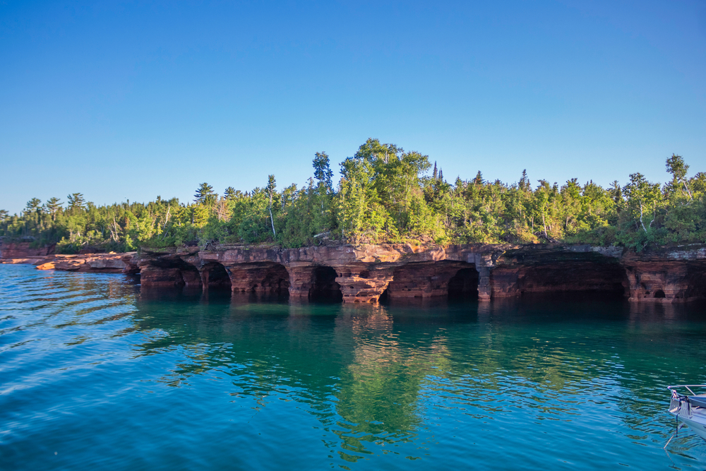 A view of the shore of one of the Apostle Islands. The shore is made up of rocky caves and cliffs with green trees growing on them. The water and the sky are both very blue. 