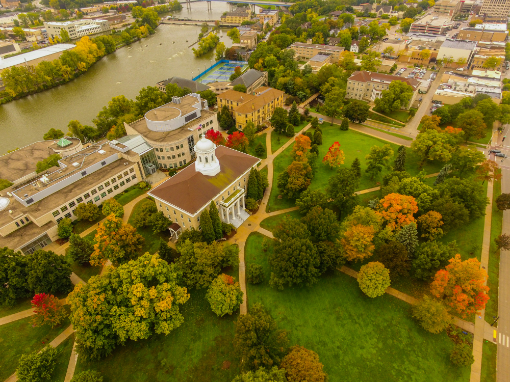 An aerial view of the college campus at Appleton. There are some modern and older buildings together on the banks of the river. At the building in the front there is a large park with green grass and lots of trees with green, red, yellow, and orange leaves. One of the best things to do in Wisconsin. 